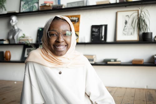 Positive African American in eyeglasses wearing traditional hijab sitting on floor and widely smiling at camera