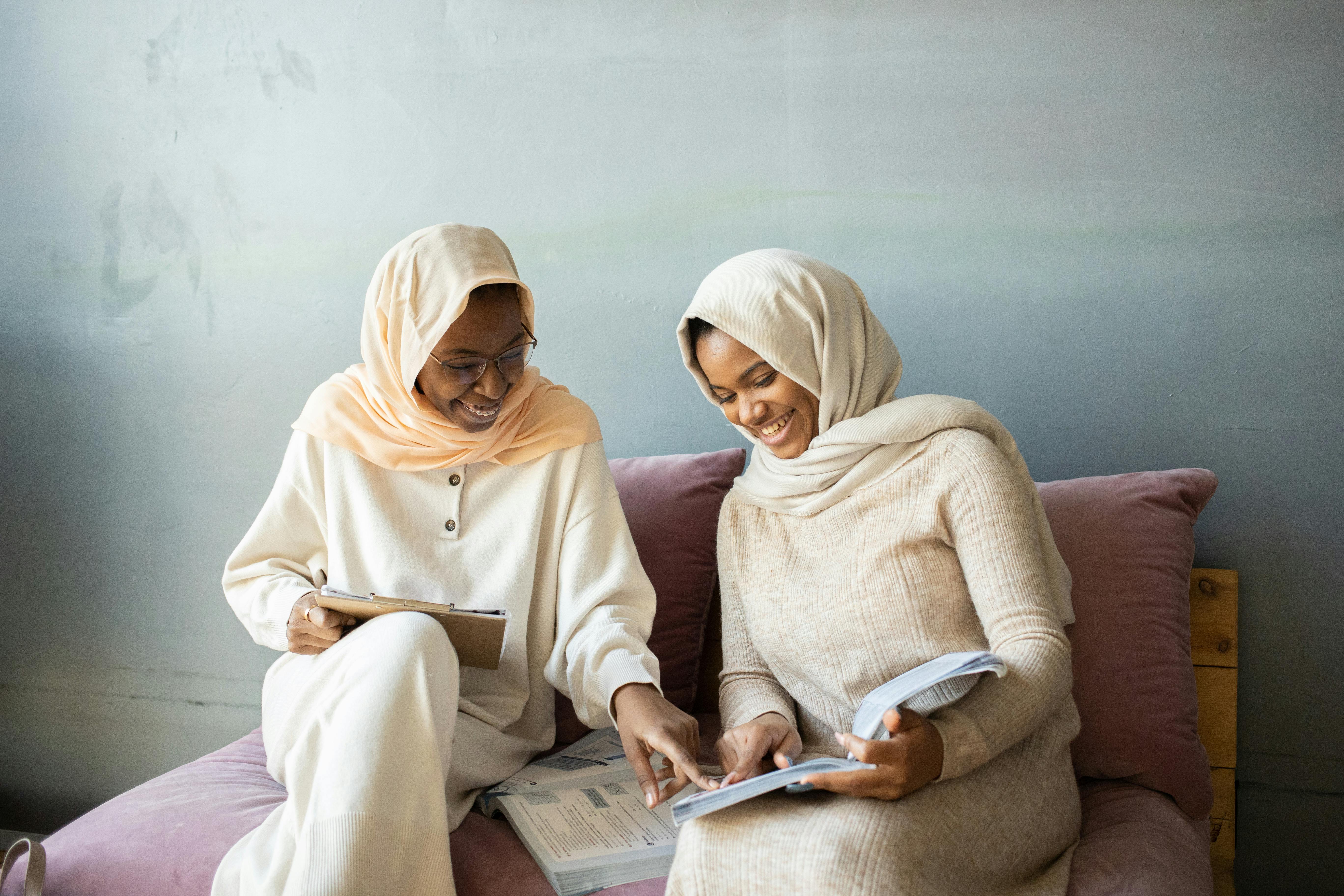 Two women in hijabs enjoy studying together indoors, sharing a moment of happiness and learning.