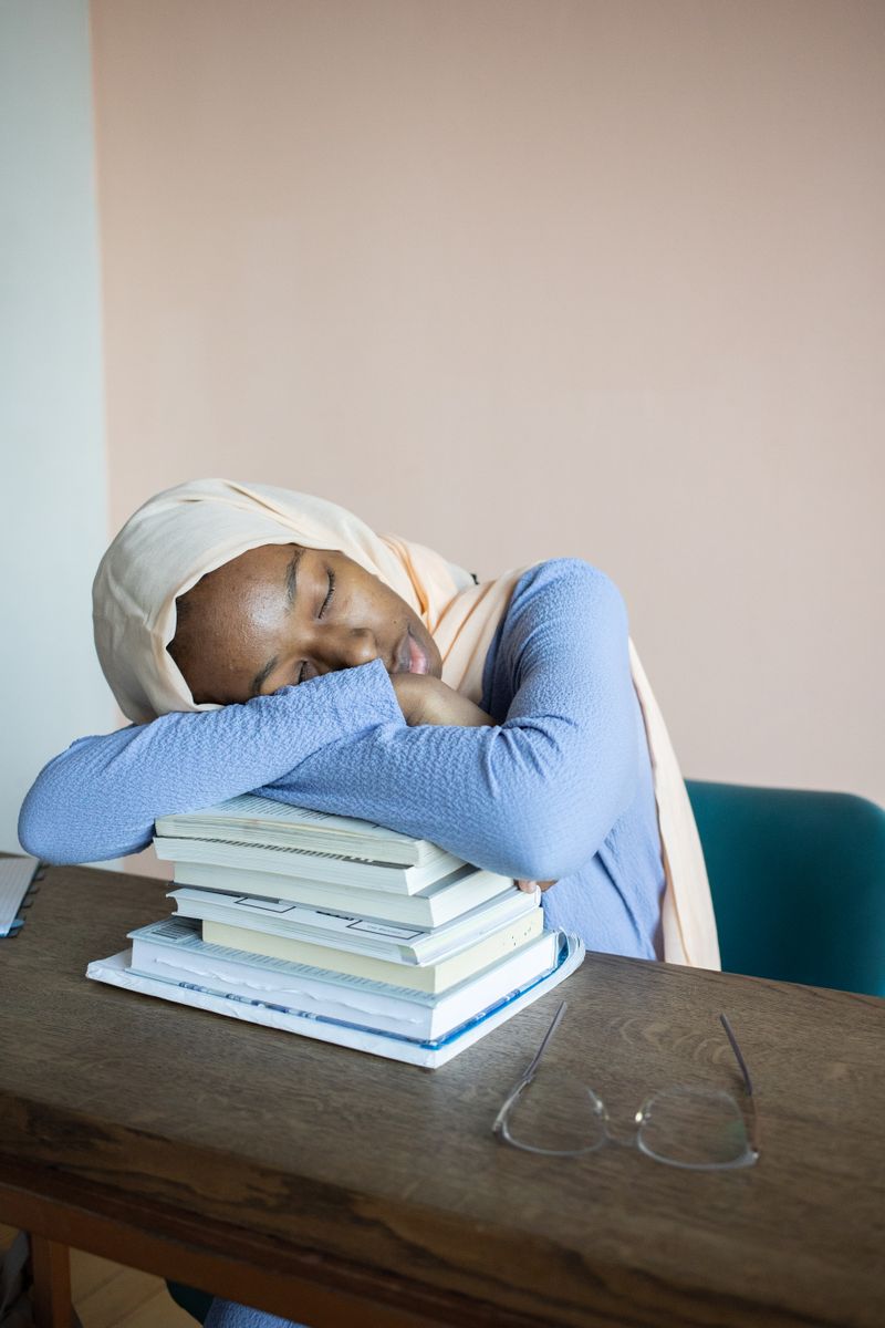 A tired Black woman in a hijab rests her head on a stack of books with her glasses set aside, an image of the deep fatigue that hypothyroidism causes and that doctors too often dismiss.