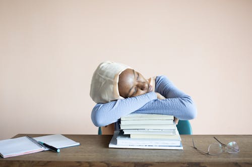 Sleeping African American female student in headscarf sitting at table with opened notebook and lying on stack of textbooks