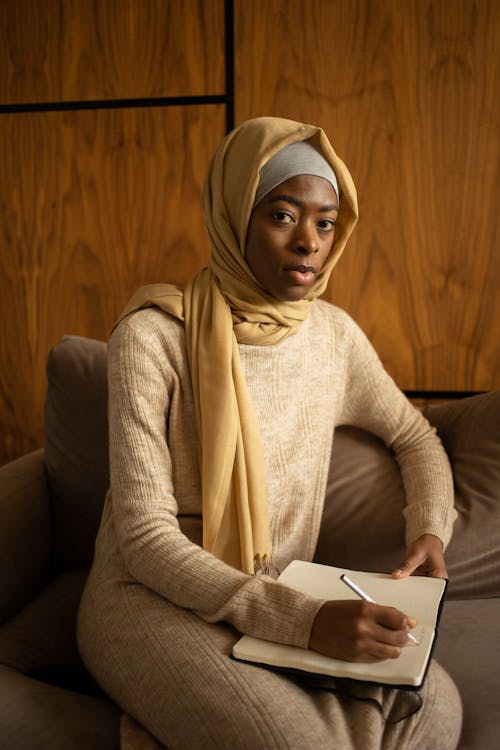 Free Concentrated African American Muslim female in hijab taking notes in planner and looking at camera while sitting on comfortable couch Stock Photo