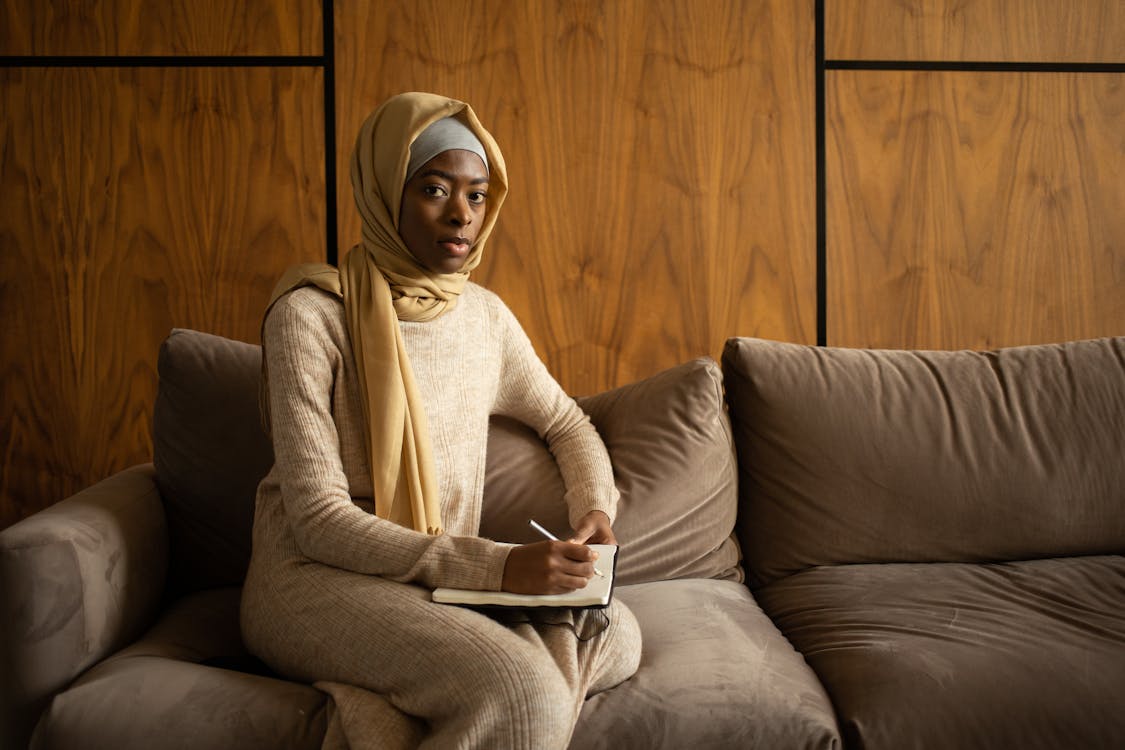 Serious black Muslim woman sitting on cozy sofa with notebook