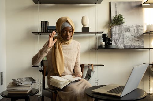 Positive Muslim African American female clerk in hijab raising arm in greeting gesture while having video call through netbook while working from home office