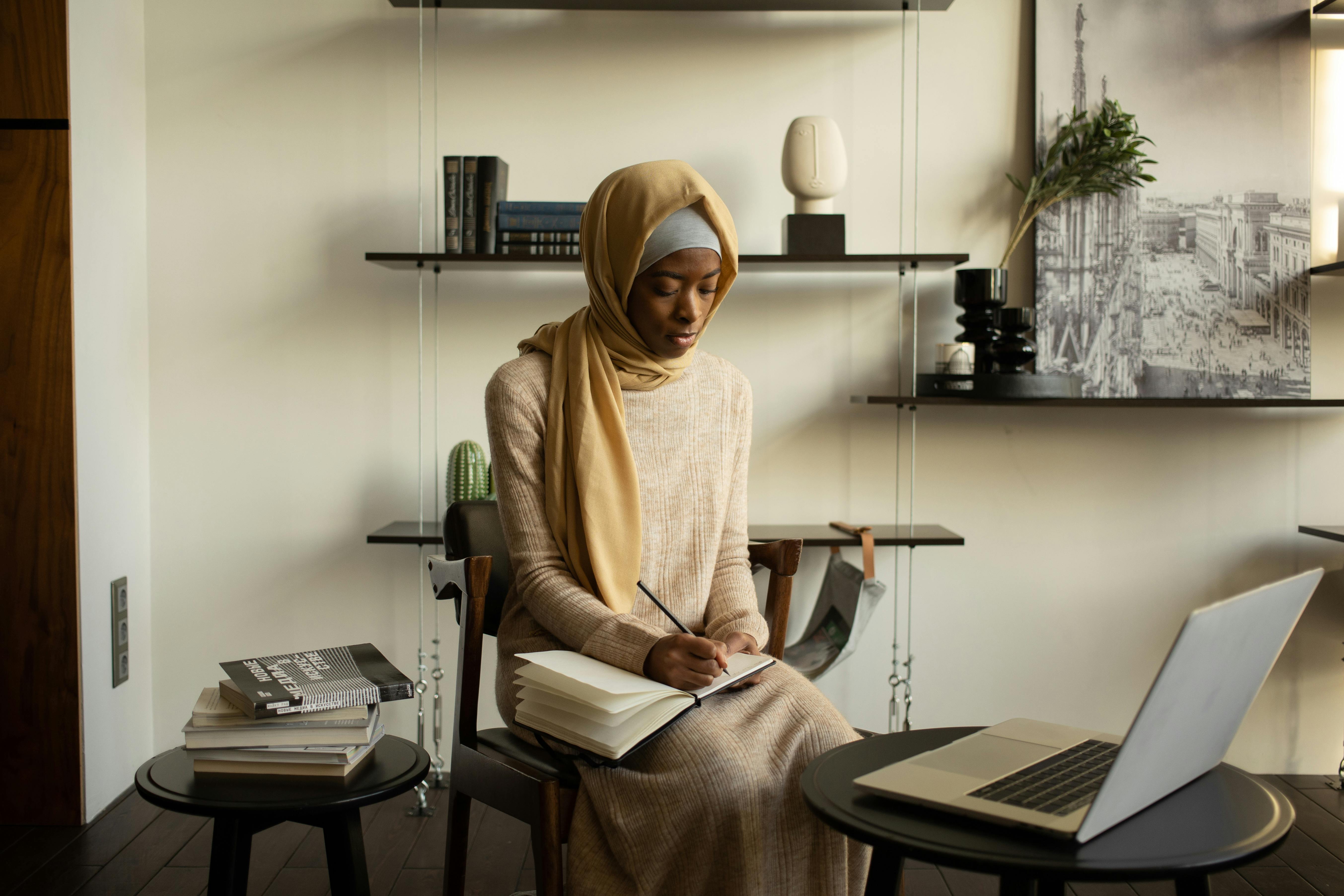 focused black muslim woman taking notes and studying via laptop