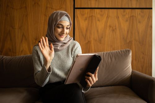 Free Cheerful young Muslim female in headscarf and sweater waving hand while having video chat via tablet while sitting on cozy sofa Stock Photo