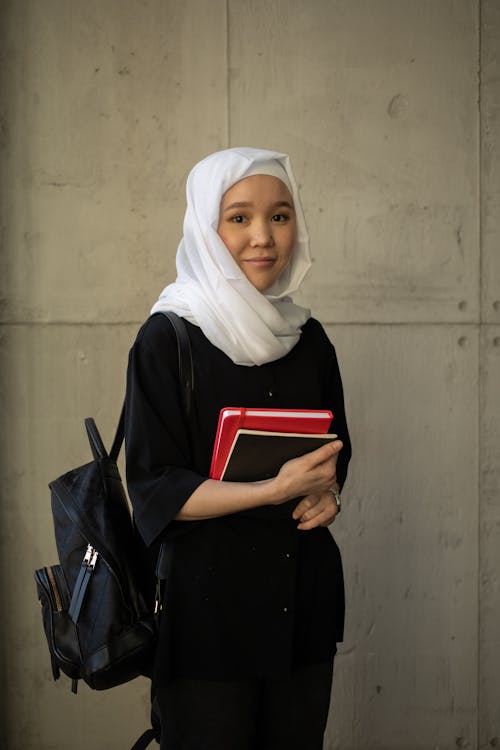 Positive young Muslim female student in black outfit and white hijab holding books and backpack while looking at camera and standing near building wall