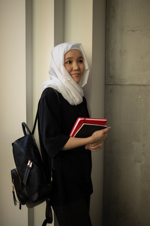 Dreamy Asian female student wearing traditional hijab looking away while standing with notepads and backpack in university campus during studies