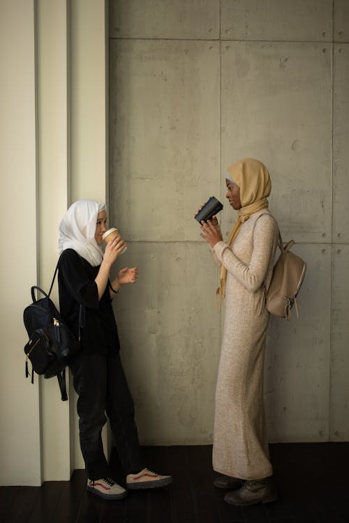 Free Full body side view of Muslim female students with backpacks drinking takeaway hot beverages and looking at each other while standing in university campus Stock Photo