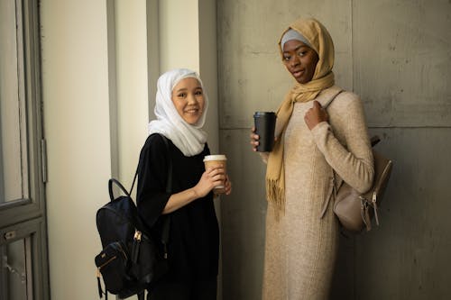 Free Positive diverse Muslim female students in traditional headscarves with backpacks and takeaway hot drinks looking at camera while standing in corridor Stock Photo