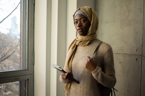 Calm Muslim female student in traditional headscarf looking away while standing with workbooks near wall in corridor of university during studies