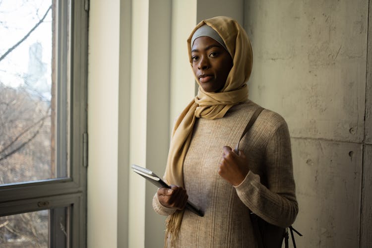 Serious Black Woman In Hijab With Notebooks Near Window