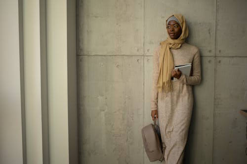 Pensive Muslim female student in traditional headscarf looking away while standing near wall with rucksack and workbooks in university campus