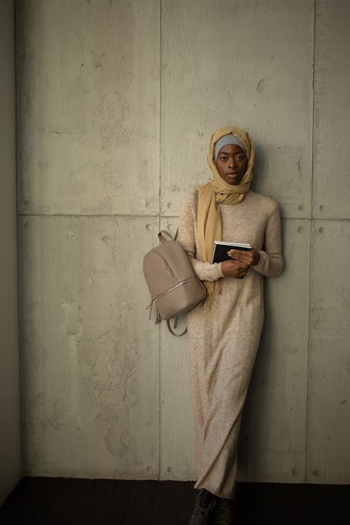 Full body of Muslim female student in traditional headscarf looking at camera while standing with backpack and notebooks in corridor of university