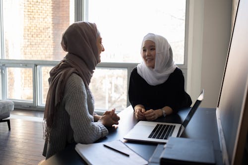 Positive multiethnic Muslim female students in headscarves looking at each other at table with netbook and copybook in classroom while studying together