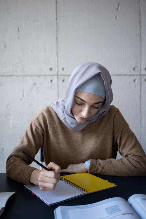 Free Concentrated Muslim female student in headscarf taking notes in workbook while sitting at table in classroom during lesson at university Stock Photo