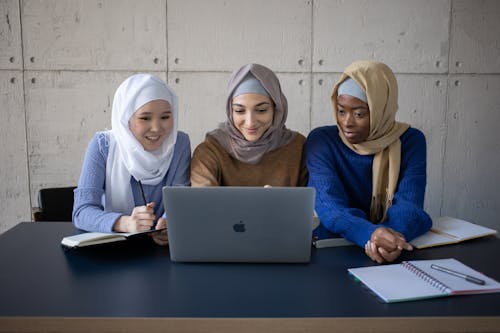 Positive multiracial women in traditional hijabs surfing modern netbook at table with copybooks while preparing for lesson together in university