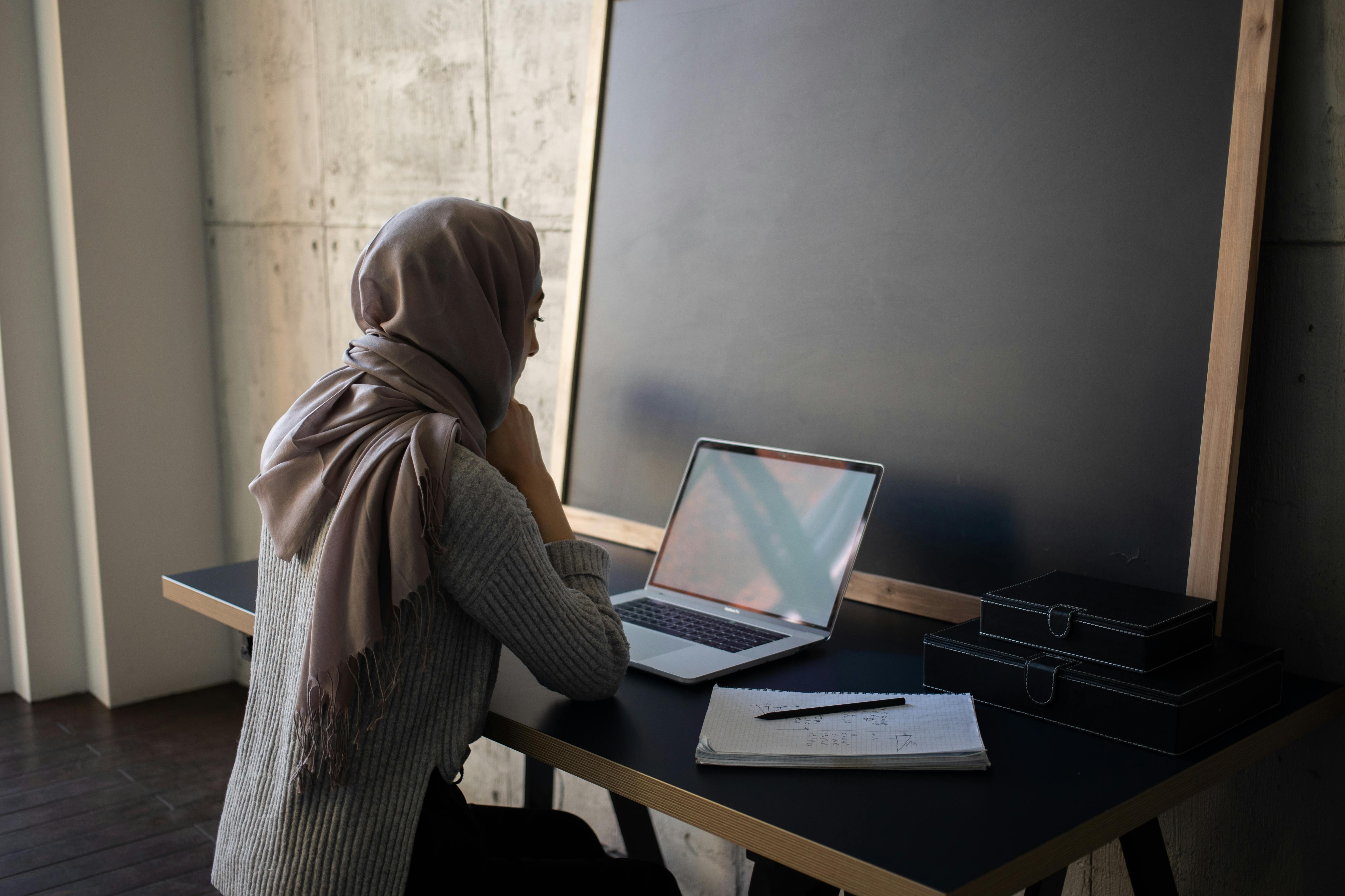 serious ethnic woman in hijab browsing laptop while doing homework