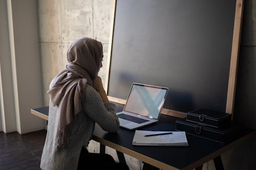 Side view of Muslim female student in traditional headscarf looking at screen of netbook while preparing for lesson an table with workbook near blackboard
