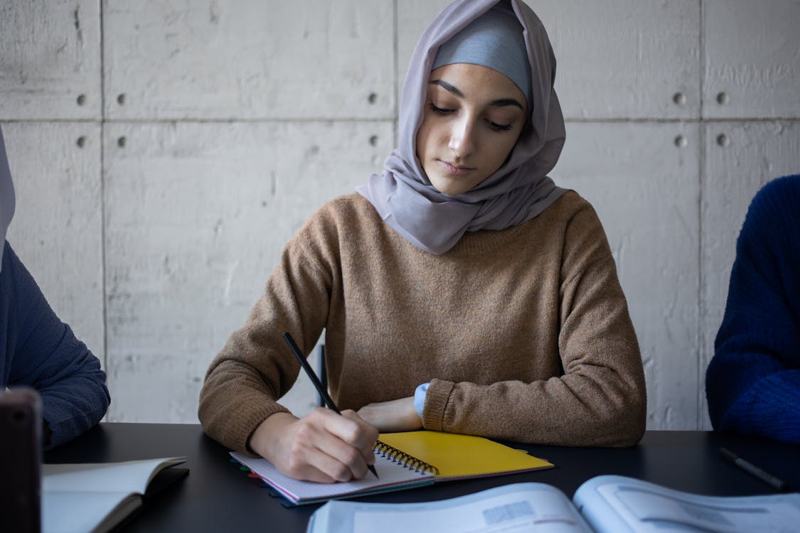 Diligent ethnic woman in hijab taking notes in workbook during lesson