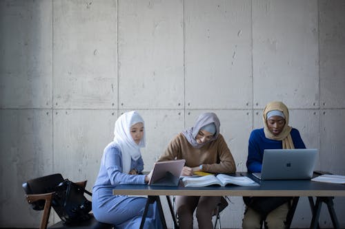 Free Concentrated multiracial female students in headscarves sitting at table while writing in copybook and browsing tablet with laptop during studies at university Stock Photo