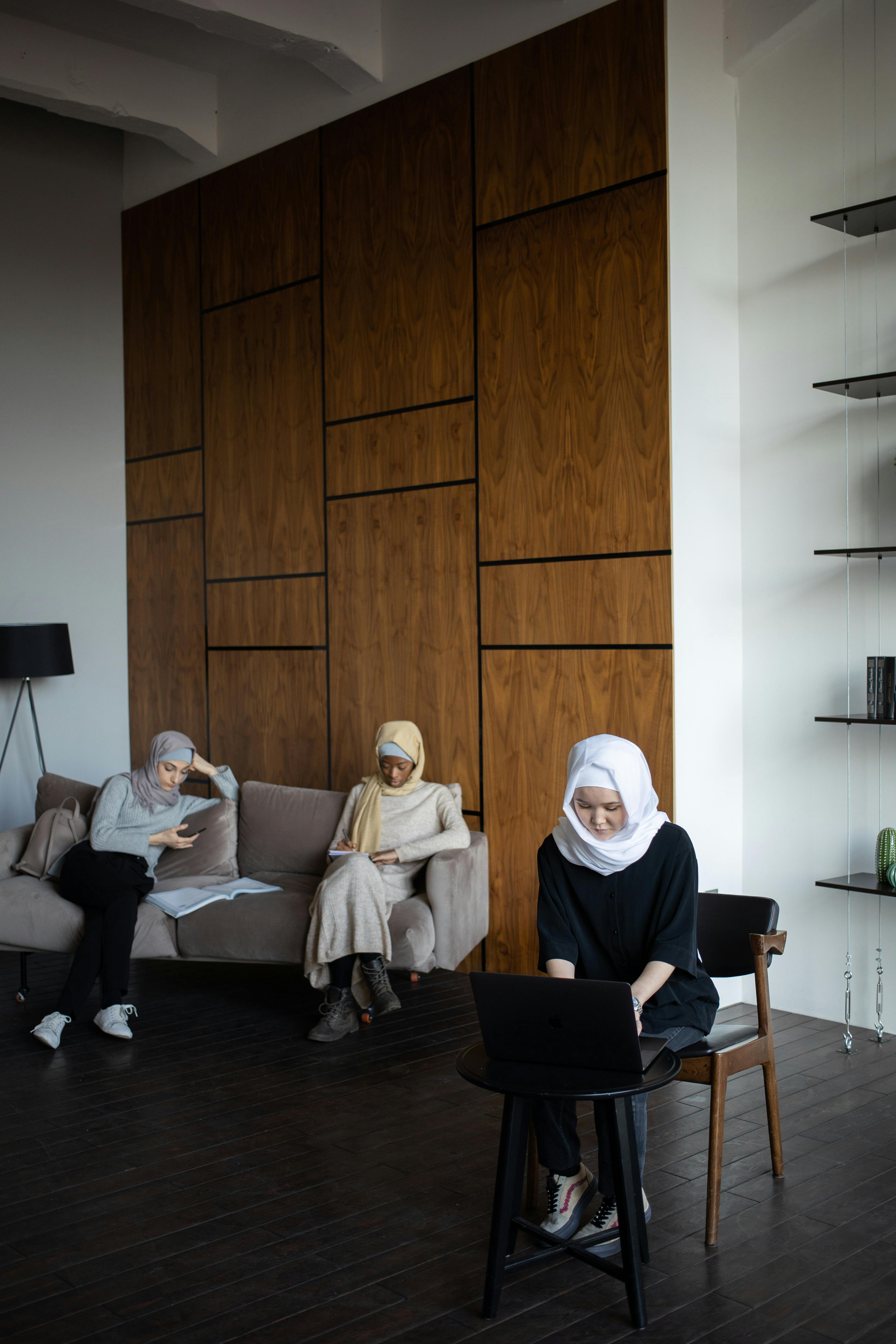 Full body of concentrated multiracial female students in traditional headscarves sitting in modern apartment and browsing modern devices while preparing for lesson together