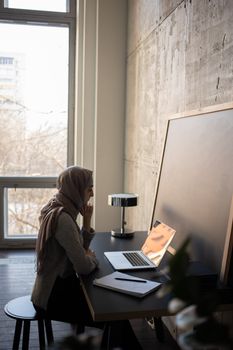 Side view of serious Muslim female student in headscarf browsing netbook at table with copybook and chalkboard while doing task for lesson