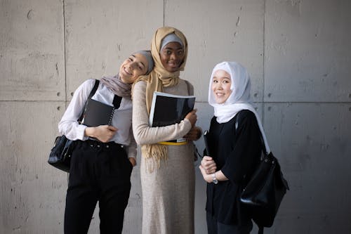 Delighted multiethnic female students in hijabs looking at camera while standing with backpacks and copybooks in hands near wall in corridor of university