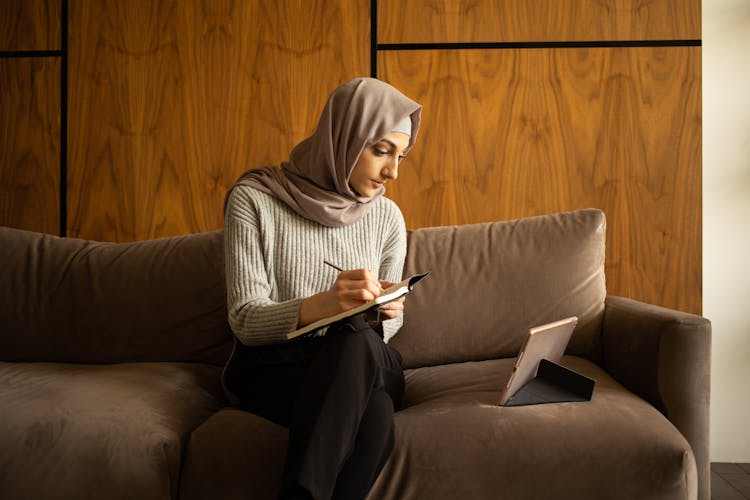 Serious Ethnic Woman In Hijab Writing In Notebook While Sitting On Couch With Tablet