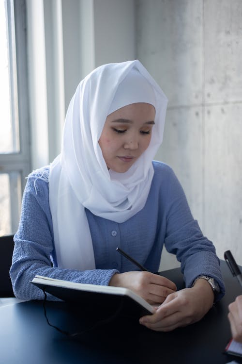 Attentive Asian woman in hijab writing in copybook during studies