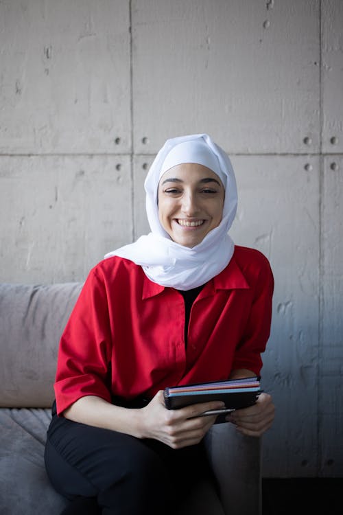 Positive Muslim female in headscarf smiling and looking at camera while sitting on sofa with copybook during studies in university