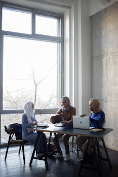Three women in hijabs collaborate and study with laptops in a bright, modern classroom.