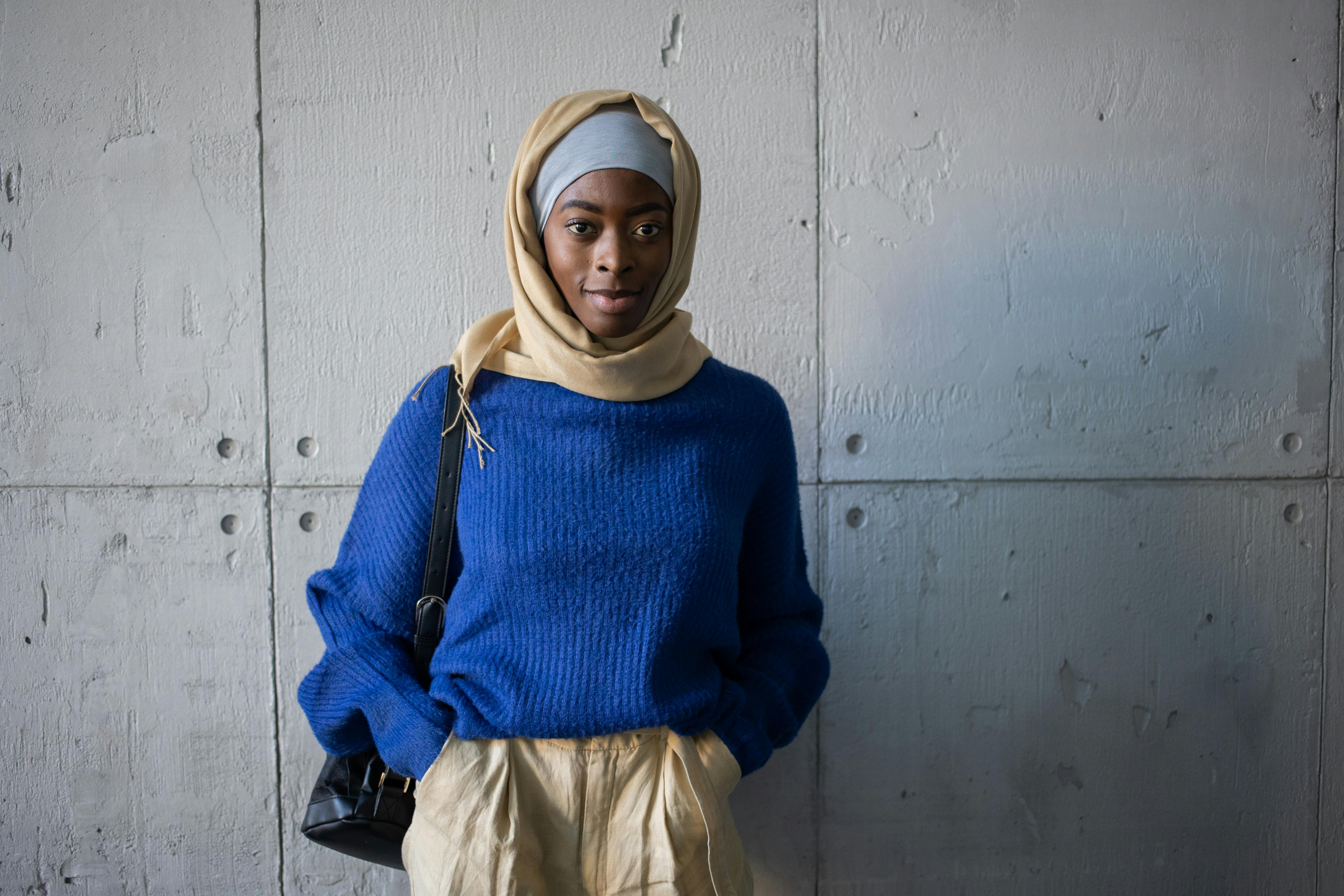 Positive Muslim female student in traditional headscarf with backpack looking at camera while standing with hands in pockets near wall in university corridor