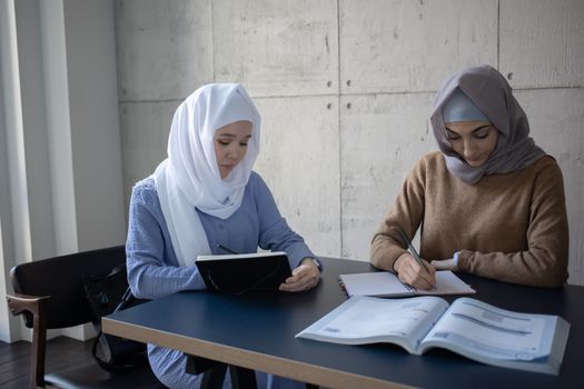 Concentrated multiracial female students in headscarves taking notes in workbooks  while sitting at table with textbook during lesson preparation in university