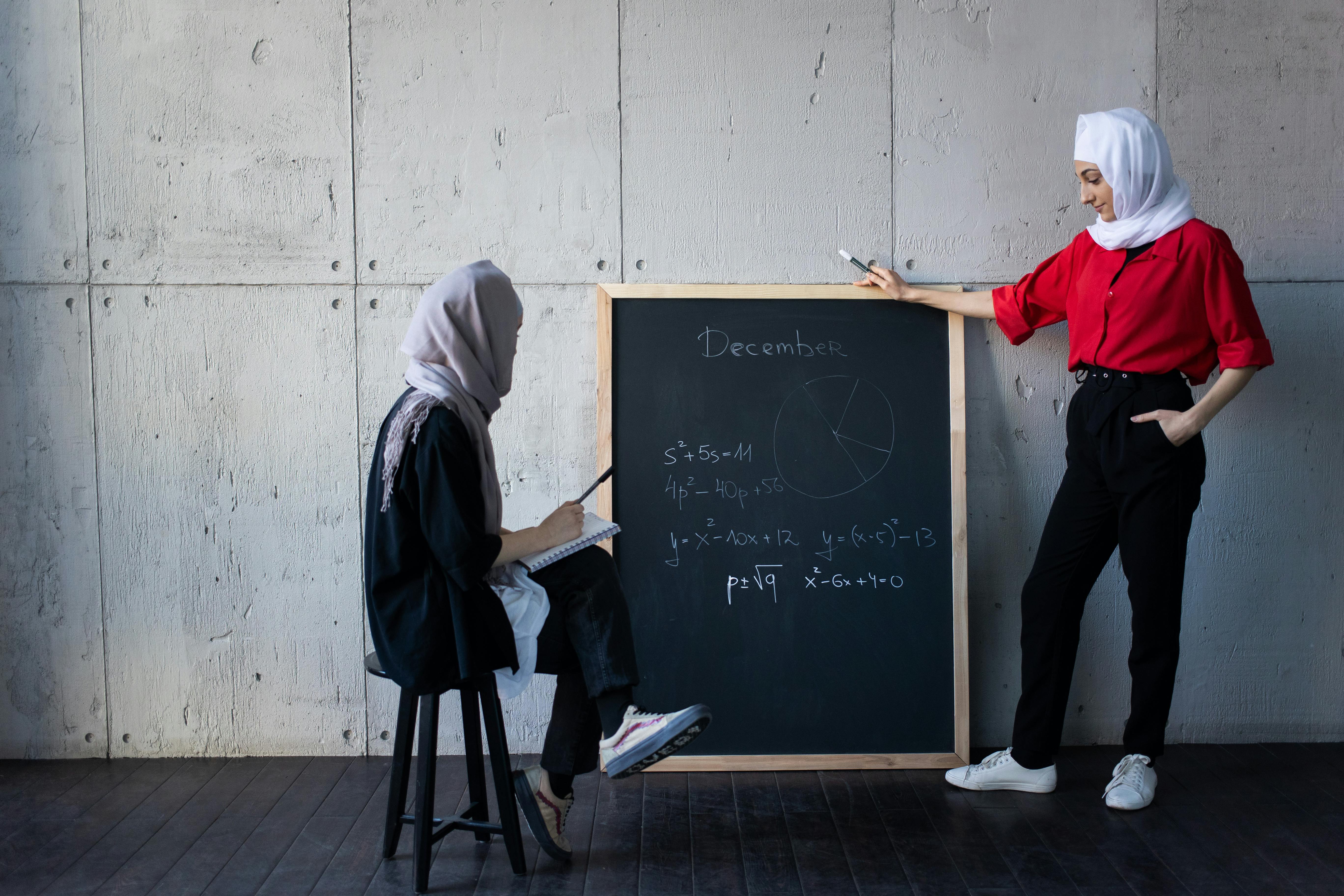Two women in hijabs sharing mathematical knowledge on a chalkboard indoors.