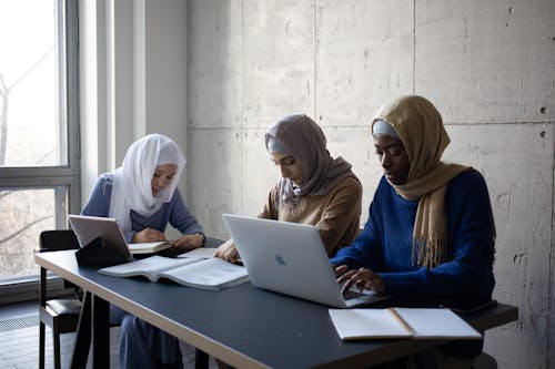 Serious young multiethnic Islamic female students in casual clothes and hijab sitting at table while surfing on netbooks and taking notes in notepads near textbook in light room near window