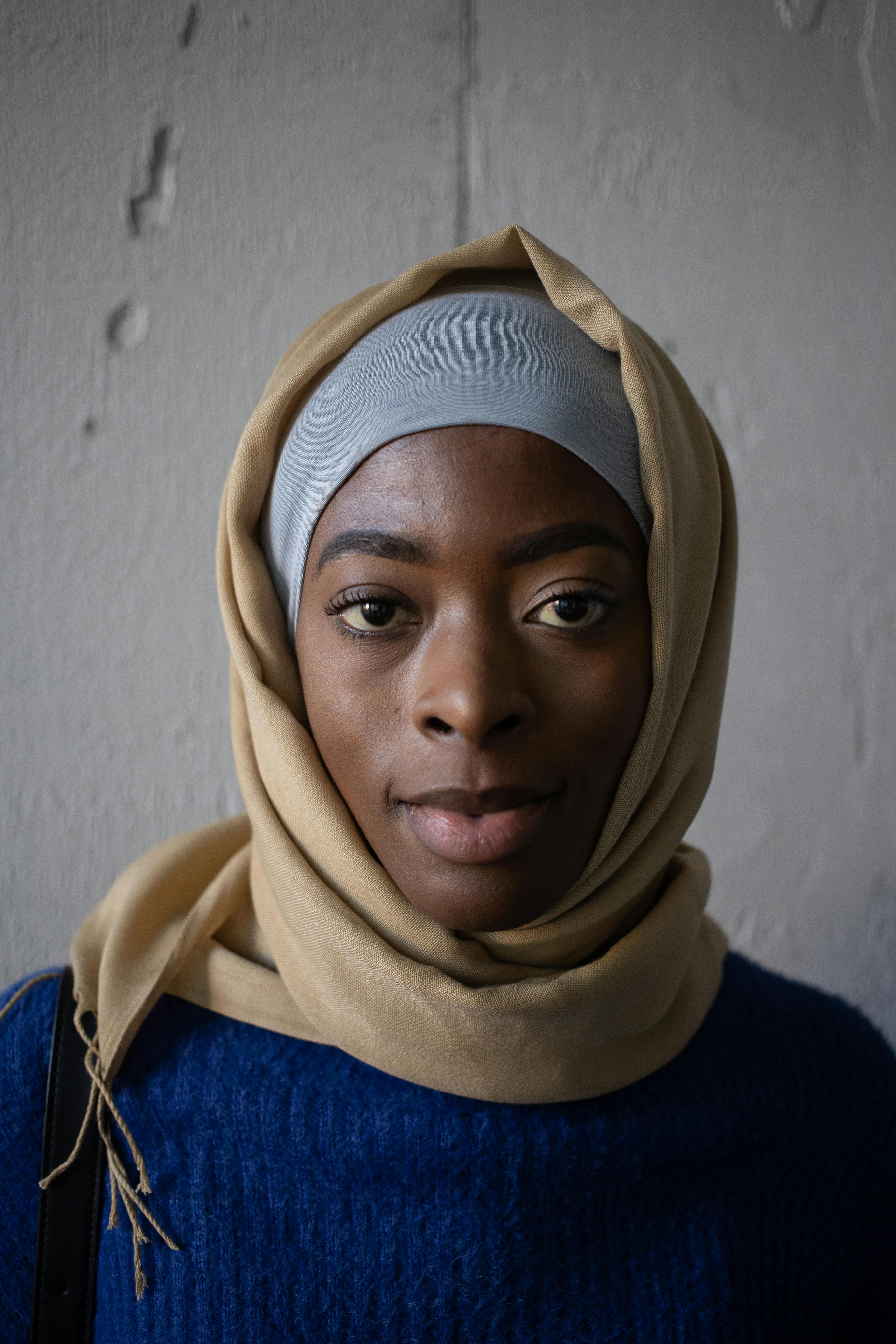 Confident black Islamic woman wearing casual clothes and hijab looking at camera near gray shabby wall in light place