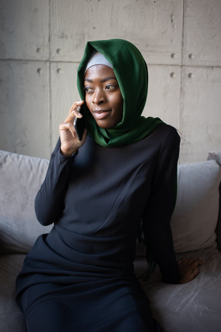 Black Muslim Lady In Hijab Speaking On Cellphone On Couch