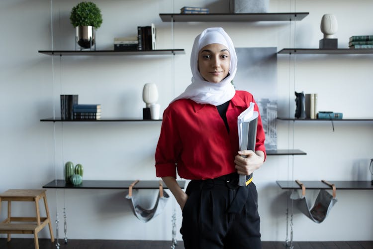 Islamic Woman With Textbooks Near Shelves With Decorations In House