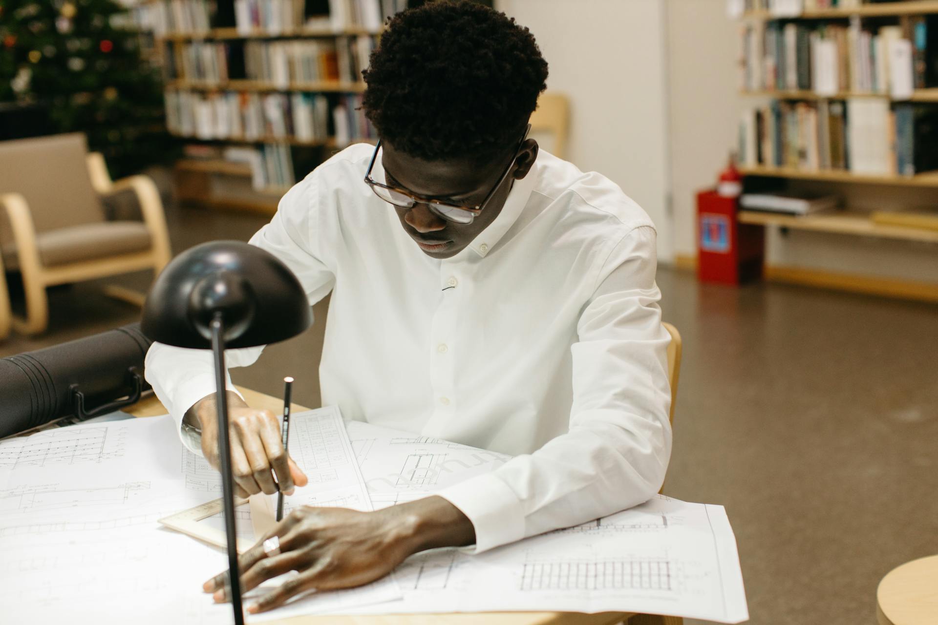 A young man intensely focused on drafting architectural plans in a library setting, showcasing concentration and work in progress.