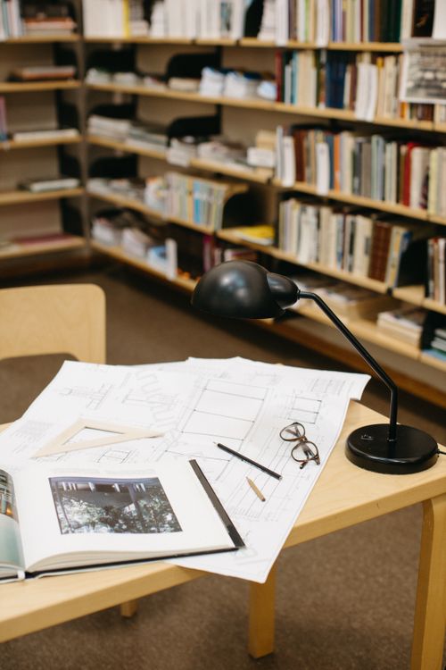 Free A tidy workspace in a library setting, featuring blueprints, a desk lamp, and bookshelves. Stock Photo