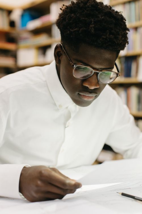 Free Young man in glasses reading and studying, surrounded by books in a library. Stock Photo