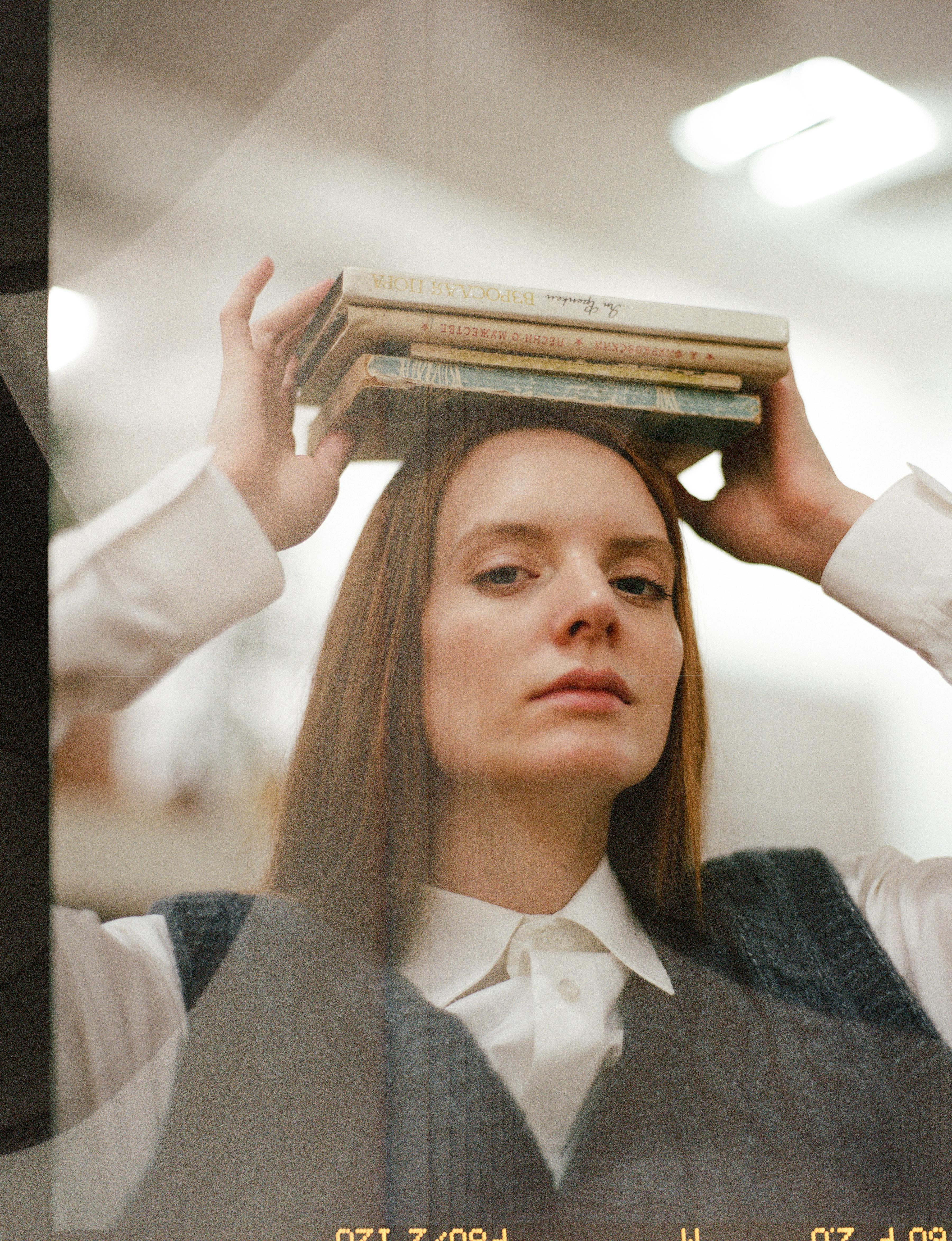 Young woman with red hair balancing books on her head in a modern indoor space.