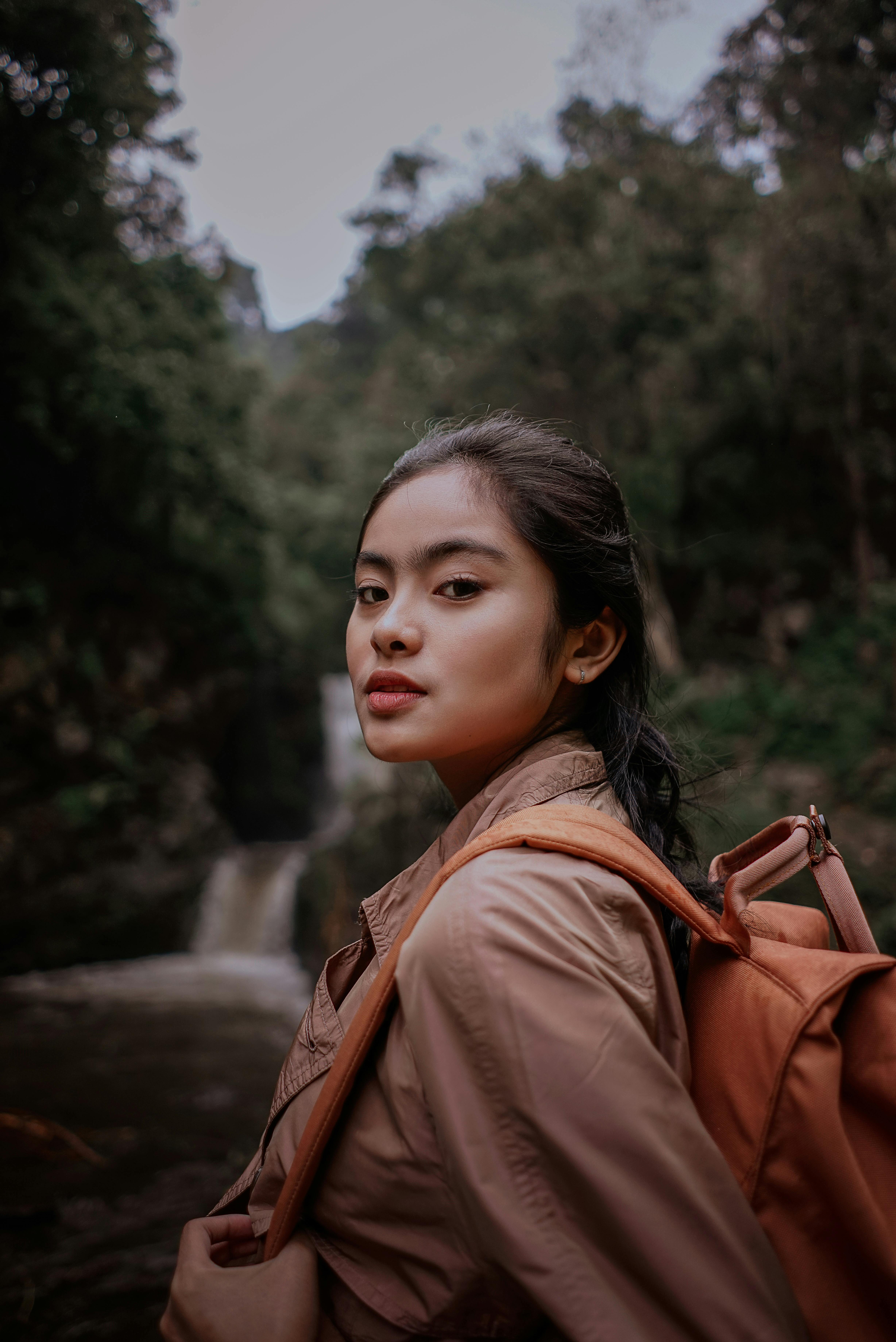 Young Asian lady in casual outfit with backpack looking at camera while standing near green forest and river with waterfall in summer day