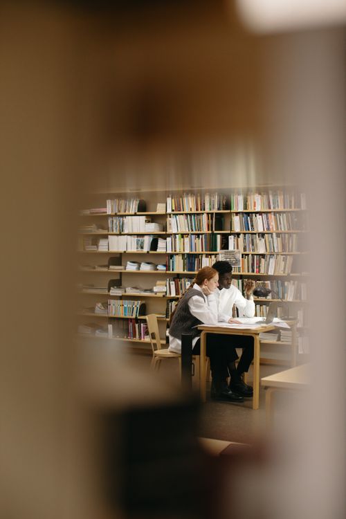 Free Two students collaborating at a library desk surrounded by bookshelves. Stock Photo