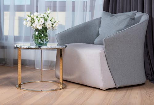 Comfortable modern styled gray armchair placed near small round marble table with vase of fresh white flowers