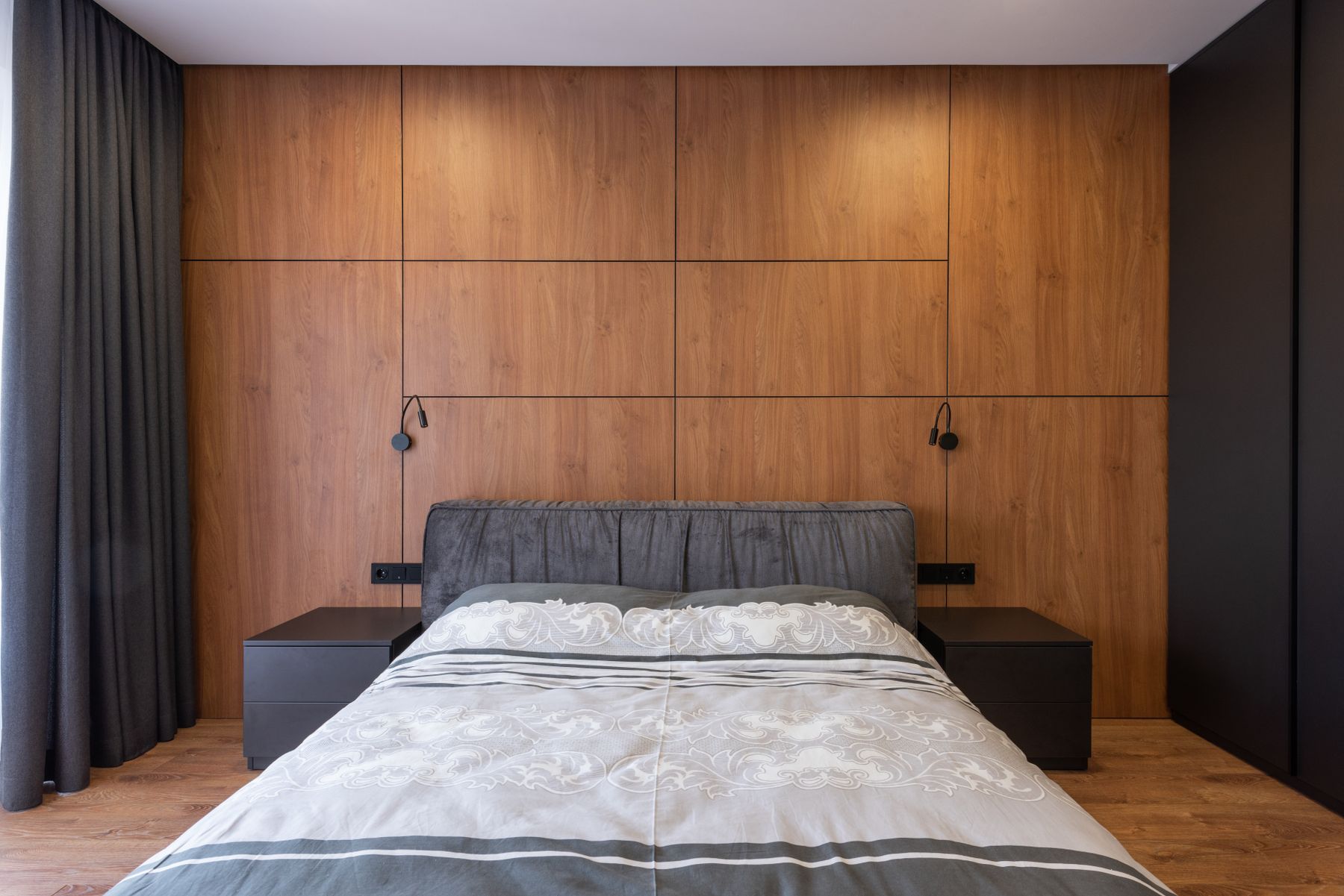 Modern bedroom with a gray upholstered bed, full wood-paneled wall, dark bedside tables and long charcoal curtains