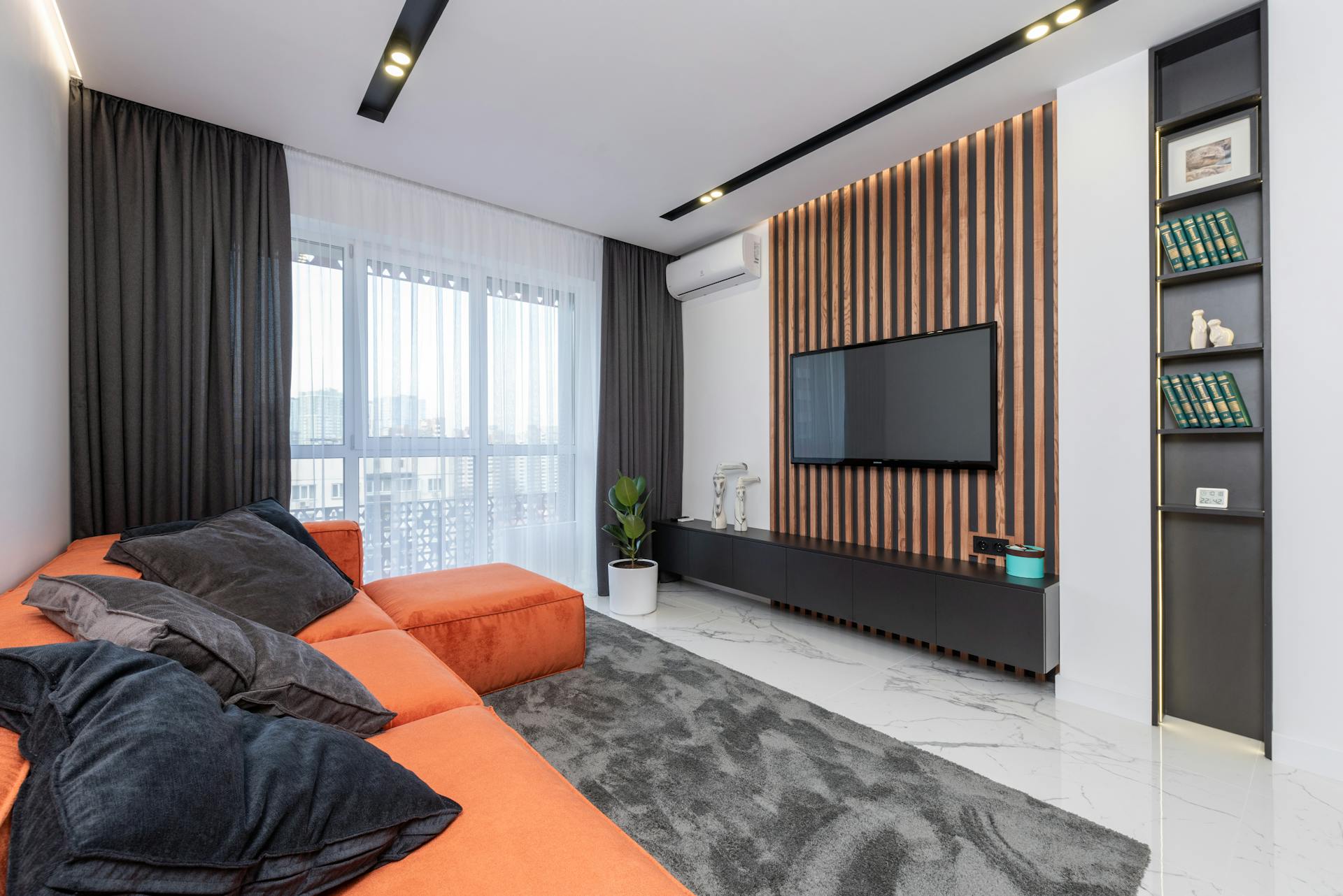 Modern spacious room with window and black cushions on orange couch placed on rug against wall with striped decorations and TV