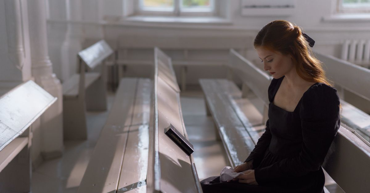 A person sitting in a sunlit, empty stone chapel with hands folded, eyes closed, surrounded by silence
