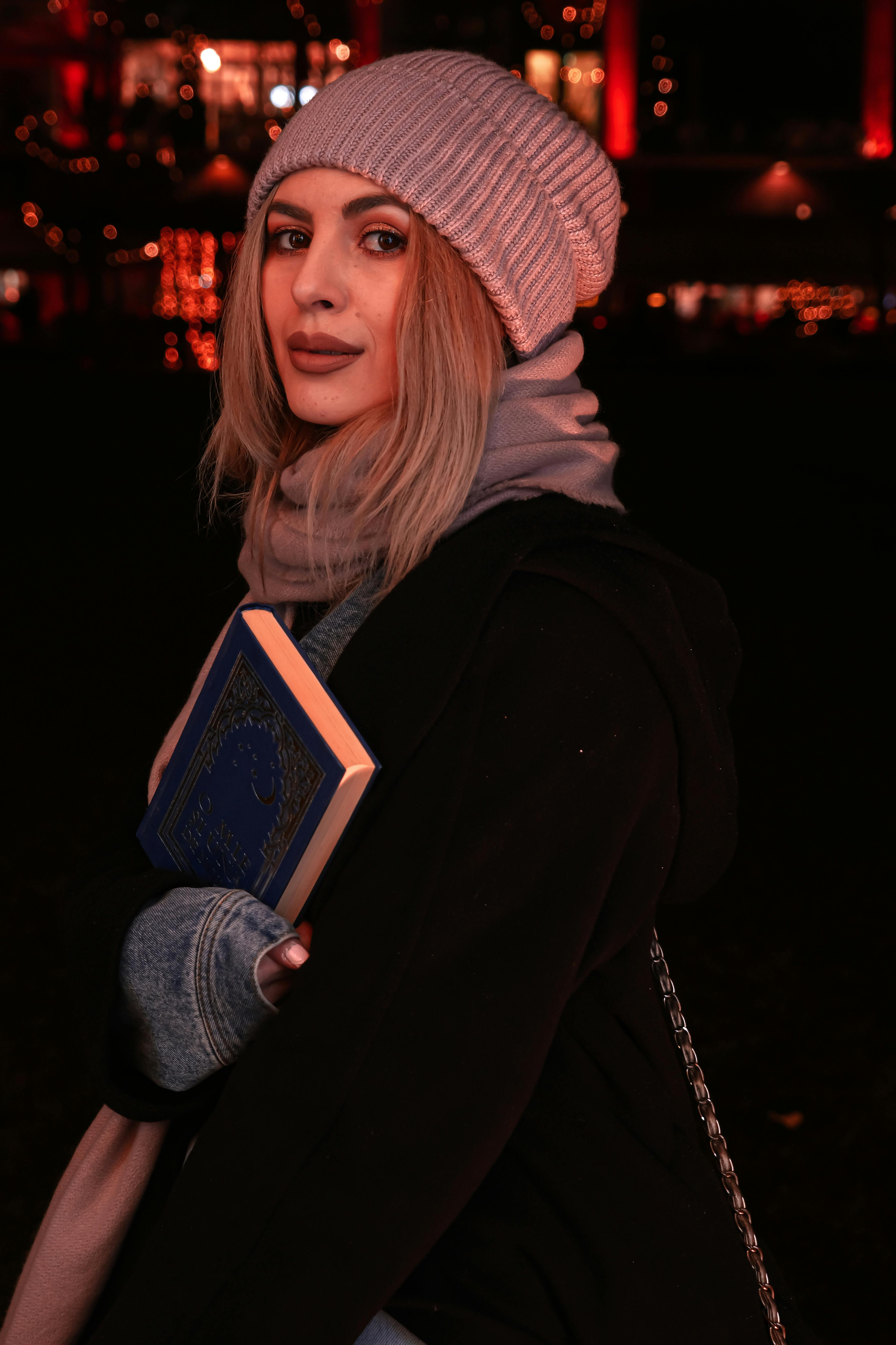 Stylish woman in winter clothing holding a book at a vibrant night market.