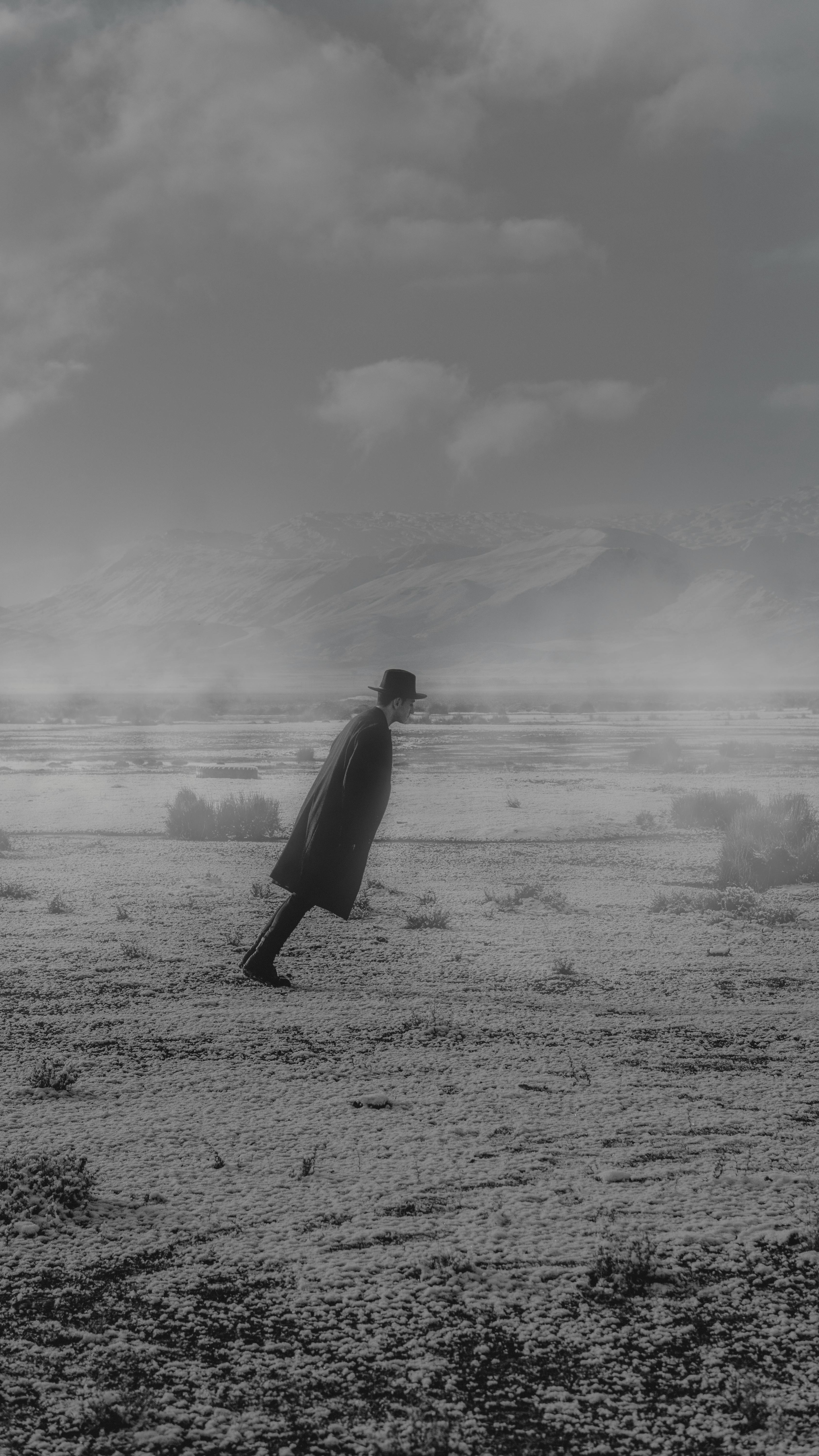 A man in a coat and hat balances in a misty, expansive wilderness landscape in black and white.